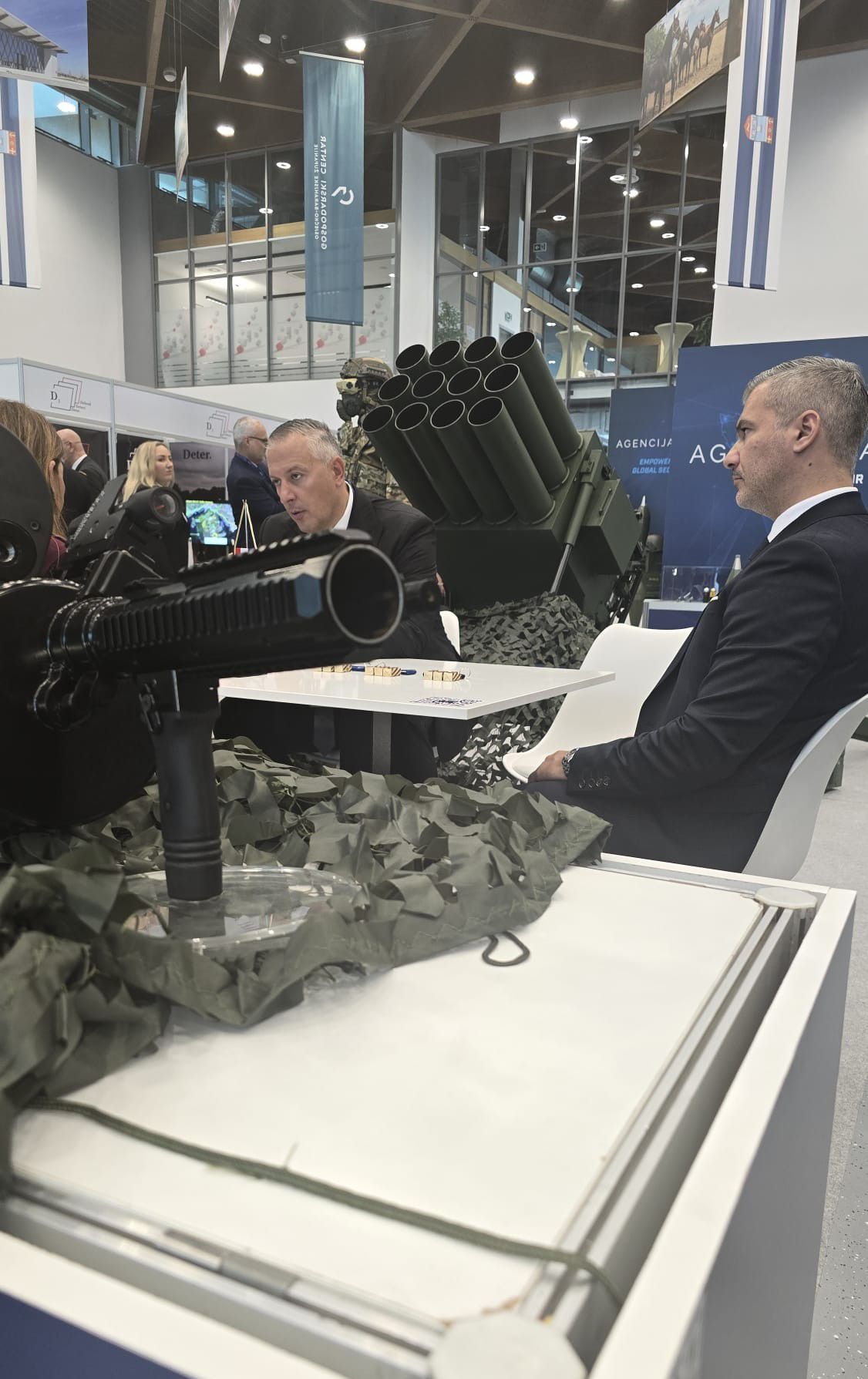 DEFCROS exhibitor meeting — defense industry executives in business discussion alongside a multiple-launch rocket system on display