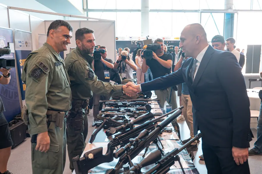 Handshake over a table of small arms at an exhibitor stand, with press cameras behind